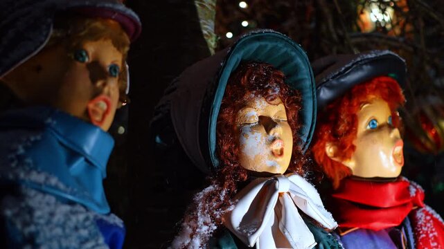 Close up faces of vintage Christmas caroler statues outdoors. Macro shot of holiday singing figures with snow on faces and winter hats under night lighting in NYC.