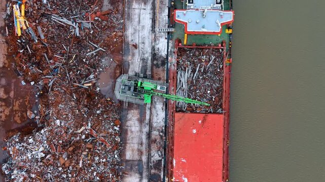 Aerial top view of crane loading scrap metal into ship. Green hydraulic material handler moving recycled metal waste into a cargo vessel hold at port.