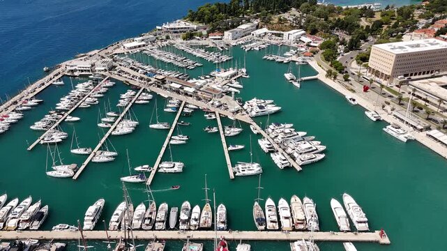 Aerial drone view over marina in Split with yachts and boats moored along piers in clear turquoise water. Harbor, docks and waterfront, sailing and marine tourism.