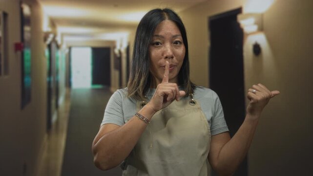 Woman wearing apron with finger to lips for silence and pointing to hallway in a building; quiet secrecy.