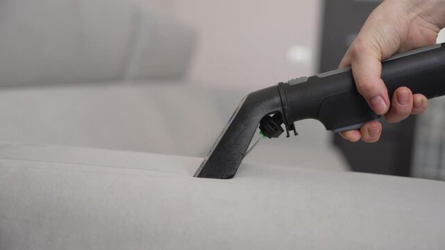 Close up of a hand using an upholstery extractor or steam cleaner nozzle to deep clean a light gray sofa. The video shows the process of removing dirt and stains from fabric furniture