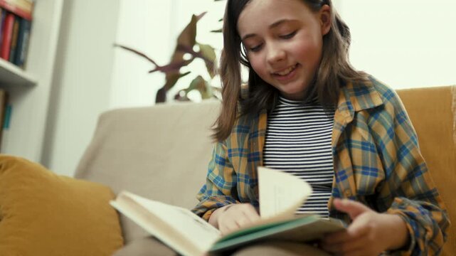 Happy preteen girl reading a book on a cozy couch at home, smiling preteen child enjoying literature in a bright living room, 4K high quality lifestyle footage.
