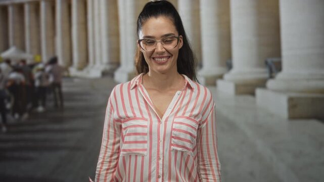 Young hispanic woman wearing glasses smiles in building colonnade among columns and walkers; friendship trust.