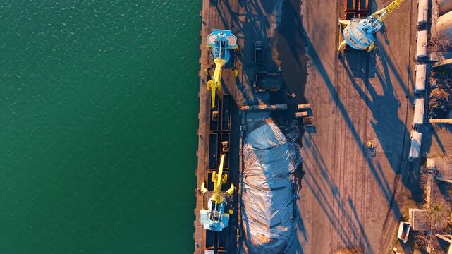 Aerial top down view of yellow cranes loading cargo ship. Industrial port scene with yellow hydraulic cranes operating near turquoise water during sunset.