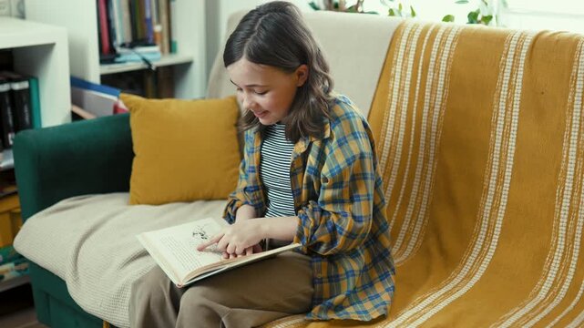 Happy preteen girl reading a book on a cozy couch at home, smiling preteen child enjoying literature in a bright living room, 4K high quality lifestyle footage.