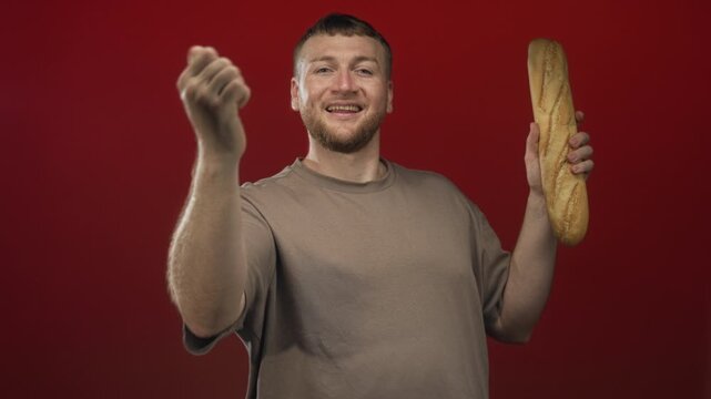 Young caucasian man holding a baguette and pointing two fingers in studio with red backdrop; confidence food humor.