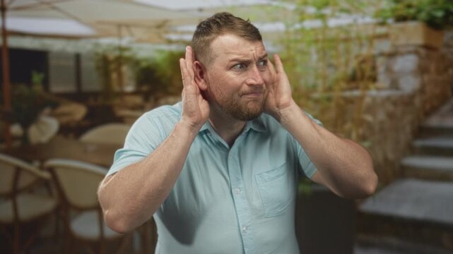 Man cups ear with hand on terrace near building entrance, young caucasian overweight with short beard and shirt sleeves, appearing strained; confusion listening.