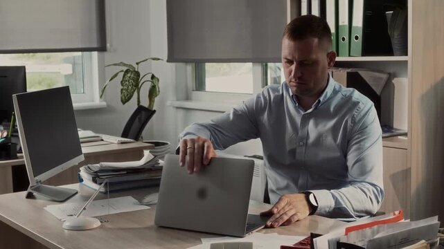 Professional office worker sits down at a desk with a compact laptop, checks the time with a wristwatch and gets to work on the computer 
