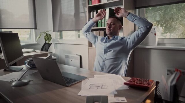 Middle-aged man leans back in his chair and puts his hands under his head to take a time out after working hard at his laptop in the office