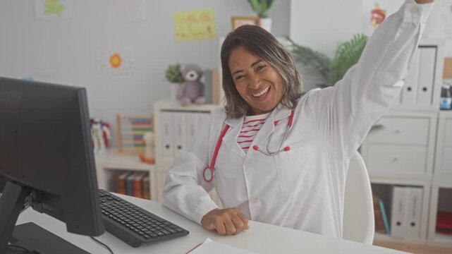 Hispanic woman doctor in white coat with stethoscope raising arm and smiling at desk with computer and pediatric toys in building; joy celebration.