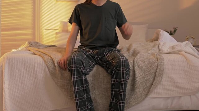 Young man with long hair in green plaid pajamas gets out of bed in the morning and puts on his house slippers 