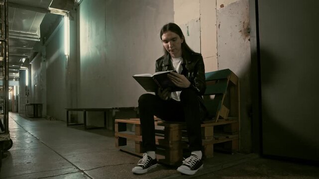 Young guy with long hair sits in a long hallway lobby corridor reading a book. Cinematic look 