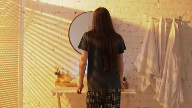 Young guy in pajamas with long brown hair goes to the sink to brush his teeth and wash his face in the early morning hours of the loft apartment