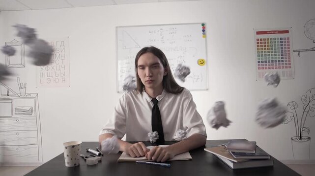 Student sitting at a desk in class is hit in the face with crumpled papers in slow motion. Bullying at school