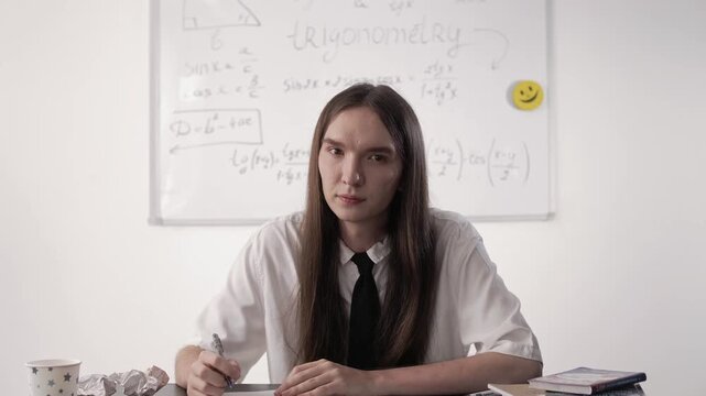Young attractive guy student with long brown hair sits at his desk during a lesson and looks intently at the camera.