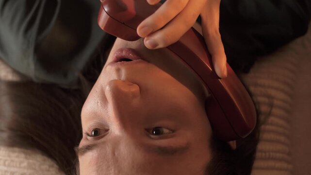 Cute young guy with long hair lying in bed singing into a telephone receiver. Portrait