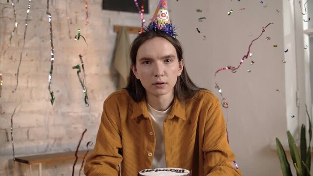 Young guy with long hair sits sad in a festive cap in flying confetti from a cracker on his birthday. Slow motion