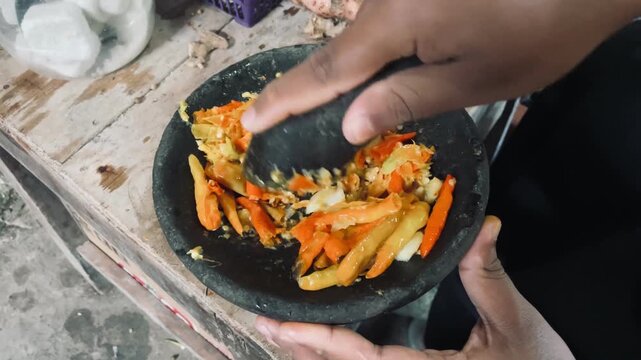 Hand grinding chili and garlic using stone mortar and pestle, capturing rustic kitchen activity, vibrant ingredients, and classic Indonesian sambal preparation.