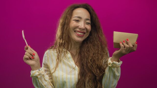 Woman smirking while holding a cardboard takeout box and plastic fork in a magenta studio; playful enjoyment.