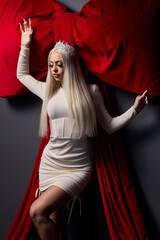 Fototapeta premium Elegant regal pose. Stunning portrait of woman with elaborate makeup and royal crown. Chic model dressed in sparkling attire standing proud against plush red drapery backdrop