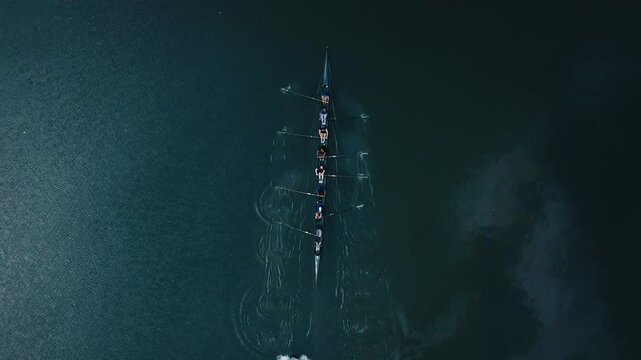 Eight person rowing crew practicing in a shell boat on a dark lake, followed by support boats