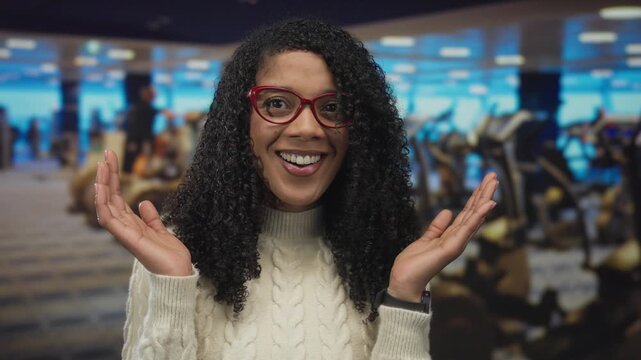 Woman wearing red glasses touches cheeks and raises hands with smile amid rows of treadmills in gymnasium; surprise.