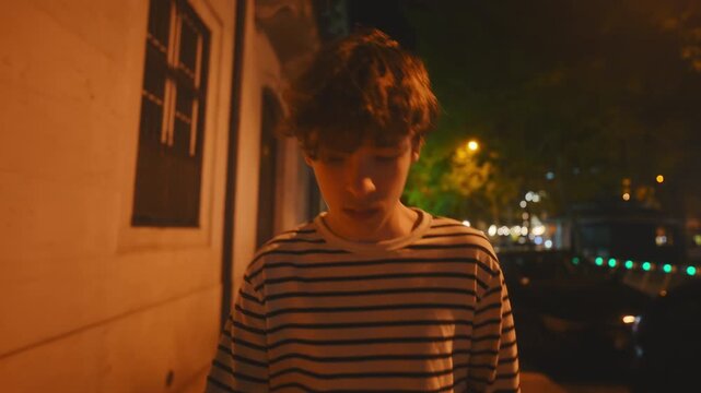 A pensive young man with curly hair walks sadly along a city sidewalk at night. The warm streetlights illuminate his downcast face as he wanders alone, lost in thought and looking troubled