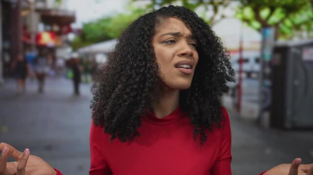 Woman with curly hair in red shirt shrugs hands on bustling street lined with storefronts; confusion doubt.