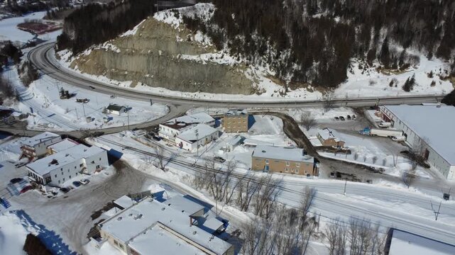 A winter drone view of Matapedia, a snow‑covered town along Route 132 at the base of a steep cliff, showing buildings, roads, railways, and a forested mountainside in Gaspesie, Quebec, Canada.