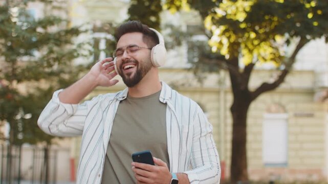 Happy Caucasian young man wearing wireless headphones choosing, listening favorite energetic music in smartphone outdoors. Smiling guy tourist walking through urban city street. Town lifestyles.