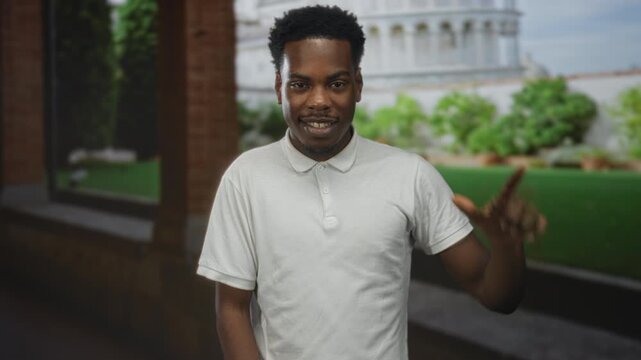 Man makes loser sign with thumb and index finger on forehead at building exterior near pisa tower under daylight; teasing.