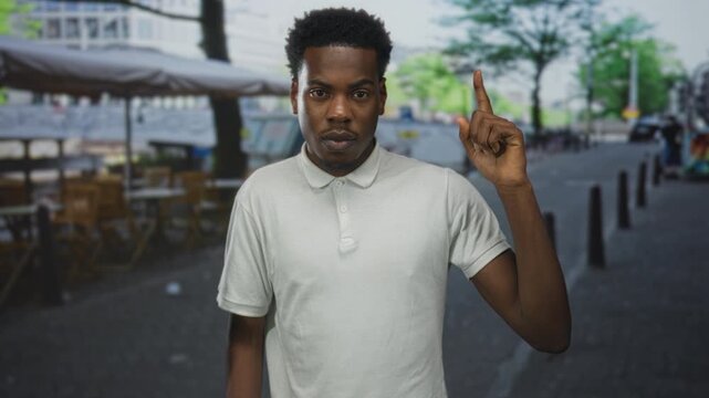Man pointing finger upward on street lined with outdoor cafe tables and city buildings behind him; attention.