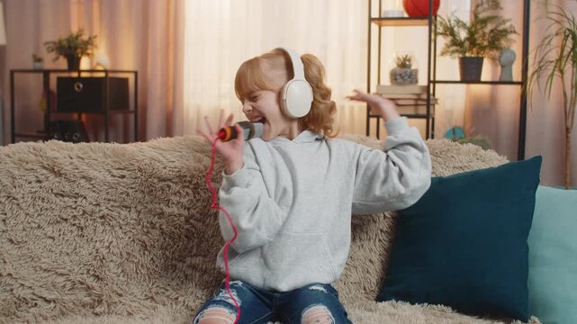 Young teenager girl wears headphones at home dancing, humming a favorite song while holding toy microphone. Blonde child kid on sofa waves hands, picturing grand performance and personal mini concert