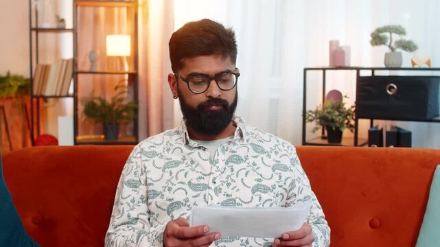 Indian man sitting at home on sofa, reading utility bills with shocked face, feeling sad about increasing prices and unpaid debt. Arabian guy reacting with stress and despair over possible bankruptcy