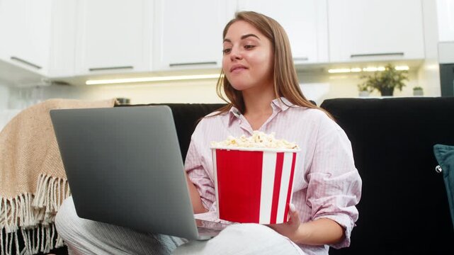 Young woman at home streams comedy on laptop eats popcorn and laughs out loud with happy mood. Caucasian girl on sofa rewinds favorite scene smiles enjoys humor comfort and small rest time and calm