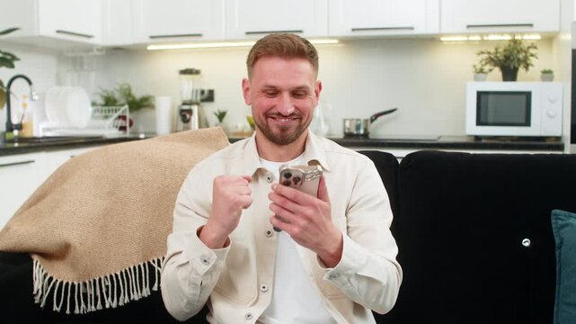 Young man at home checks smartphone sees app win good news and celebrates with big shout smile. Caucasian guy on sofa laughs holds mobile phone raises fist shows joy victory success after notification