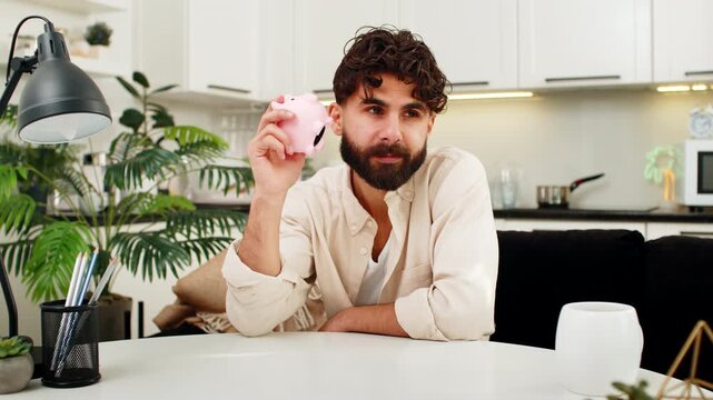 Indian man at home sits with small piggy bank in hands drops last coin inside dreams about savings. Arabian guy sitting at table shakes bank near ear gives sad smile thinks about future money goals.