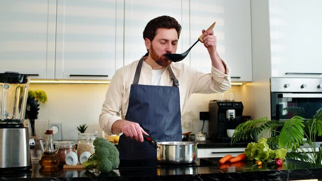 Middle-aged man adds pepper to warm soup in pot on electric stove for healthy diet dinner at home. Young guy smiles cooking fresh homemade food in kitchen indoors aiming for wellness and weight loss.