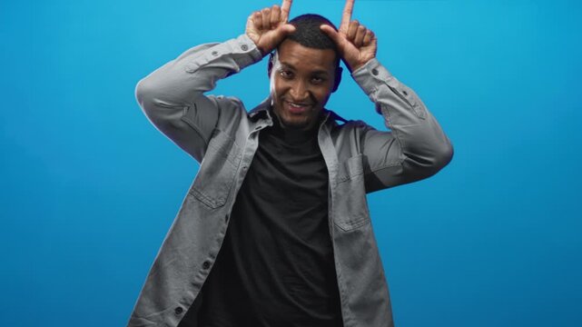 Man making horn gesture with hands and smiling against a studio blue backdrop; playful upbeat confidence.