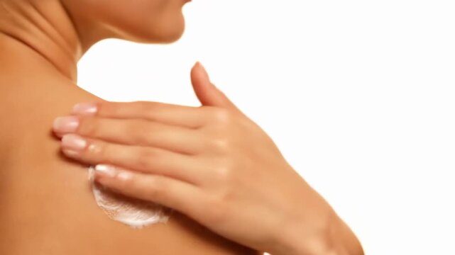 Close-up side view of a woman applying white moisturizing lotion to her shoulder and upper back against a clean white background. Skincare and beauty concept.