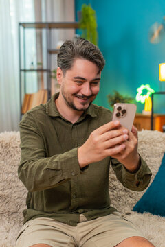Middle-aged man at home reads smartphone message smiles nodding with approval and joyful success. Young adult guy on sofa holds mobile phone enjoys positive notification showing cheery big confidence