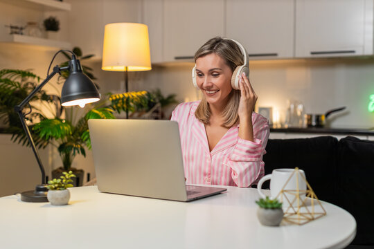 Adult woman at night home works in design tool on laptop with headphones moves layers tweaks layout smiles. Caucasian girl at table on sofa at evening nods music applies edits saves version progress.