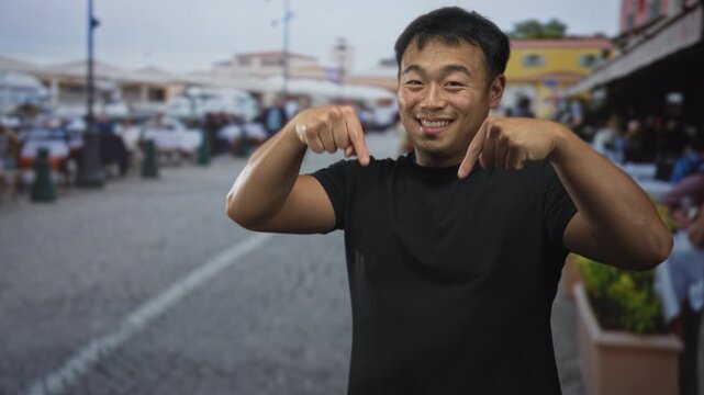Young man points fingers down at restaurant terrace on cobblestone street with a welcoming grin; enthusiasm.