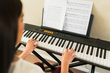 Young woman learning to play keyboard, practicing digital piano with sheet music in home setting