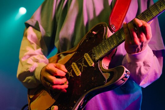 Close-up of hands playing an electric guitar with red strap and worn body finish under colorful stage lights, with haze and a spotlight in the background.