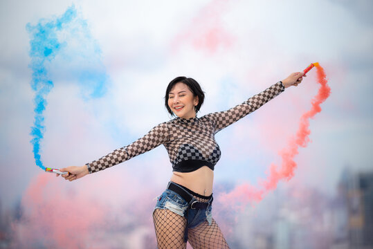 Happy woman smoke bomb rooftop urban festival blue smoke red smoke checkered top denim short fishnet tights joyful pose on city rooftop with colorful blue and red smoke swirling around her