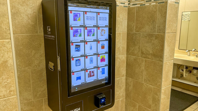 Women's washroom vending machine displaying various products on a digital screen, located in a tiled restroom environment with sinks visible in the background