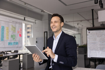 Fototapeta premium Future focused inspiration. Motivated optimistic mid adult businessman look forward with happy smile distracted from using pad in modern office workspace visualize project progress achieving success