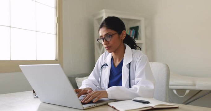 Serious young physician woman in glasses and lab coat working at computer in medical office, turning look at window, touching chin, thinking, typing on laptop, keeping electronic records of patients