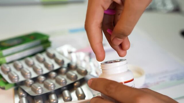 Close-up of hands opening a white pill bottle with blister packs of tablets on a desk, suggesting medication use, healthcare, and pharmacy concept.